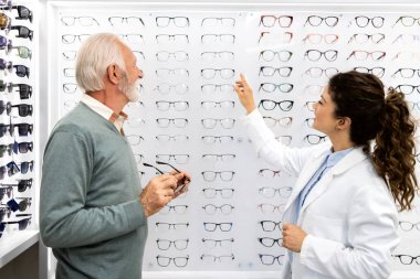 Kadın optometrist kıdemli hastaya optik mağazasından uygun bir çift gözlük seçmesini tavsiye ediyor..