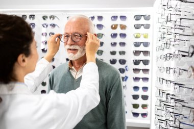 Optik mağazasında deneyimli kadın gözlükçünün yardımıyla yeni gözlük deneyen kıdemli bir hasta..