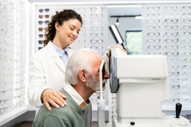 Tecrübeli optometrist yaşlı adamın görme yeteneğini kontrol ediyor. Oto kırıcı optik cihaz kullanarak odaklanıyor ve diopter kullanıyor..