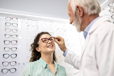Güzel, gülümseyen esmer kadın gözlük deniyor gözlükçüde tecrübeli bir optometristin yardımıyla.