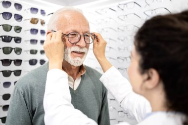 Optik mağazasında deneyimli kadın gözlükçünün yardımıyla yeni gözlük deneyen kıdemli bir hasta..