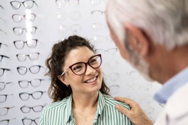 Güzel, gülümseyen esmer kadın gözlük deniyor gözlükçüde tecrübeli bir optometristin yardımıyla.