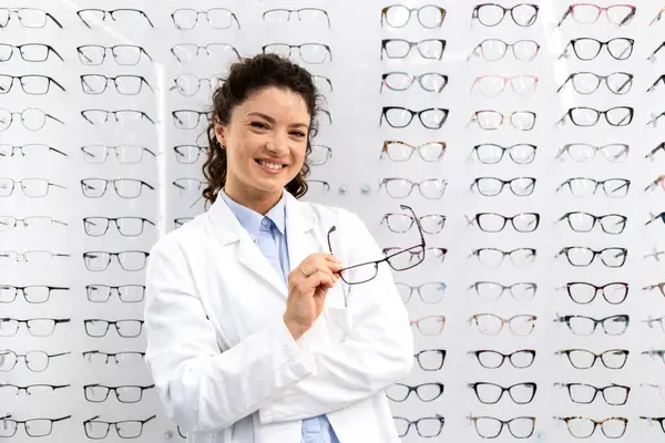 Optik mağazasında gözlük panelinin önünde duran güzel, gülümseyen kadın gözlükçünün portresi.
