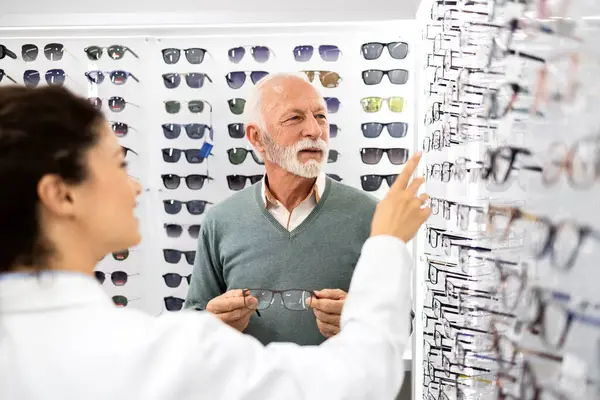 Tecrübeli bayan gözlükçü, optik mağazada yeni gözlük için mükemmel çerçeveyi seçmesine yardım ediyor..