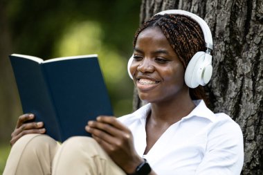 Doğada kitap okuyan ve müzik dinlerken dinlenen güzel Afrikalı genç bir kadının yakın görüntüsü.