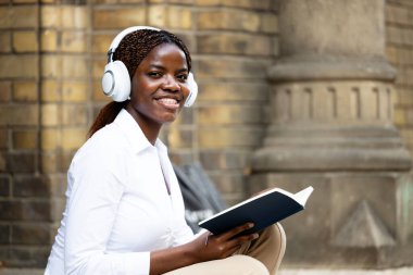 Genç Afrikalı kız öğrenci üniversitenin önünde kitap okuyor ve dinleniyor..
