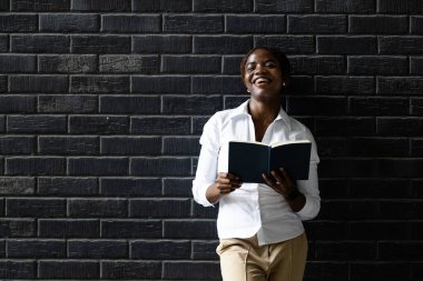 Duvara yaslanmış ve elinde kitap tutan zarif siyah Afrikalı kadın portresi..