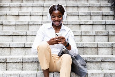 Güzel siyah Afrikalı kadın müzik dinliyor ve sokakta akıllı telefon kullanıyor..