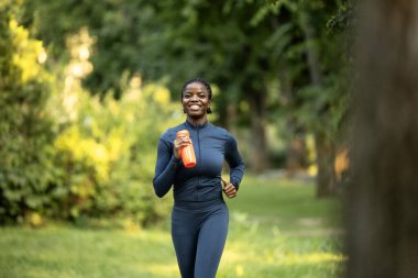 Spor elbiseli siyahi kadın dışarıda antrenman yaparken kendini iyi hissediyor. Gülümseyen siyah kadın su şişesiyle dışarıda koşuyor..