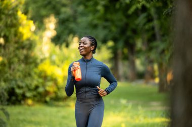 Elinde su şişesi olan Afro-Amerikalı bir kadın parkta koşuyor..