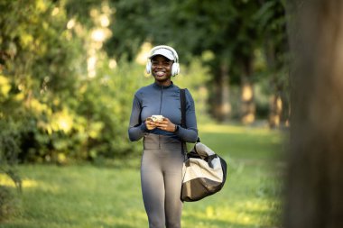 Spor çantalı zenci bayan sporcu parkta yürüyor ve akıllı telefon kullanıyor..