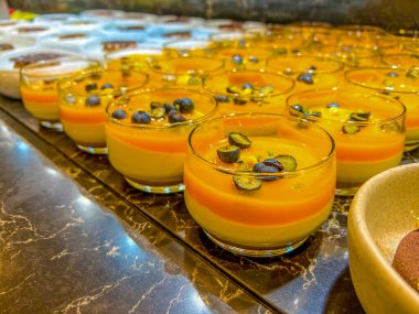 Böğürtlenlerle süslenmiş, koyu bir yüzeyde duran cam kaselerde duran kokulu mango pudingi ya da mus. Bir şenlik organizasyonunda, bir partide ya da düğünde. Açık büfe