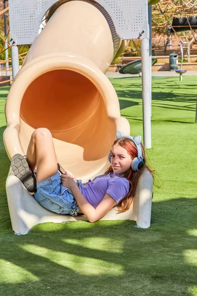 A cute, beautiful teenage girl in blue headphones with cat ears lies across a children\'s slide in a children\'s playground and listens to music on a sunny warm day