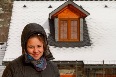 Güzel bir kadın karla kaplı çatısı olan bir evin önünde duruyor.