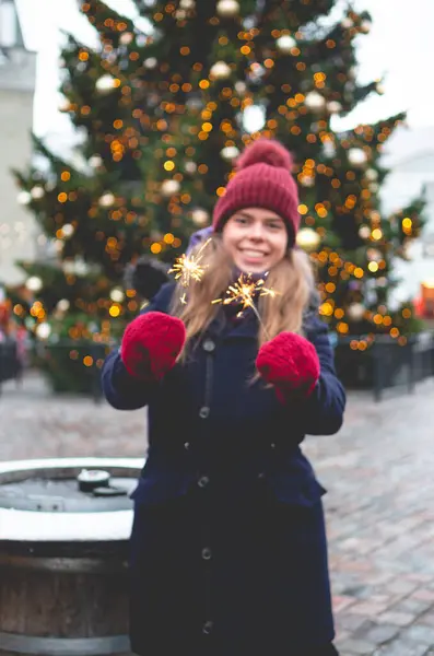 若いハンサムな笑顔の女性は輝きを保持し 背景にクリスマスツリーで寒い冬の日にタリンタウンホール広場に立っています ストック写真