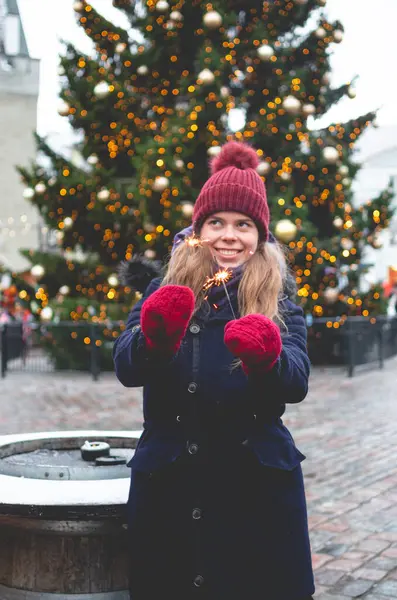 若いハンサムな笑顔の女性は輝きを保持し 背景にクリスマスツリーで寒い冬の日にタリンタウンホール広場に立っています ストック画像