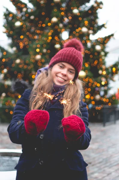 若いハンサムな笑顔の女性は輝きを保持し 背景にクリスマスツリーで寒い冬の日にタリンタウンホール広場に立っています ロイヤリティフリーのストック画像
