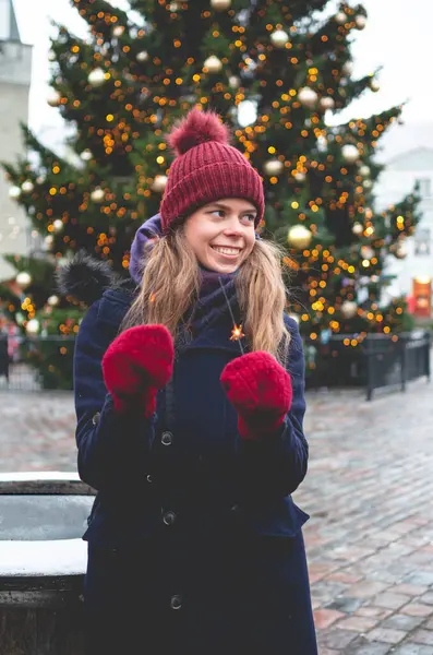 若いハンサムな笑顔の女性は輝きを保持し 背景にクリスマスツリーで寒い冬の日にタリンタウンホール広場に立っています ストック画像