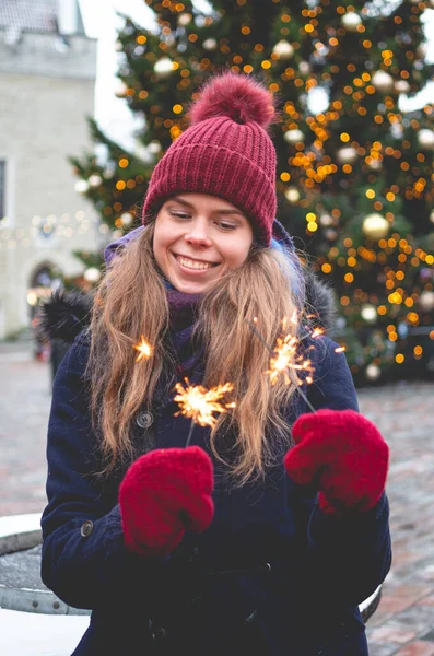 若いハンサムな笑顔の女性は輝きを保持し 背景にクリスマスツリーで寒い冬の日にタリンタウンホール広場に立っています ストックフォト