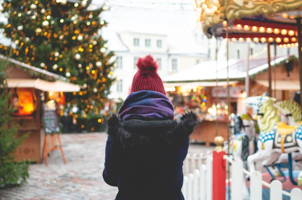 青い冬のコートを着た若い女性と クリスマスツリーとバックグラウンドのクリスマス運賃で寒い冬の日にタリンタウンホール広場に立っている赤い帽子 ストック画像