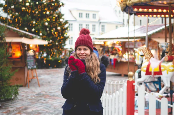 青い冬のコートにハンサムな若い女性 赤い帽子と手を一緒に握り クリスマスツリーとバックグラウンドのクリスマス運賃で寒い冬の日にタリンタウンホール広場に立って ストック写真
