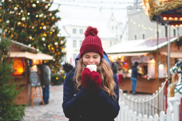青い冬のコート 赤い帽子 手袋のハンサムな若い女性はコカを楽しみ クリスマスツリーとバックグラウンドのクリスマス運賃で寒い冬の日にタリンタウンホール広場に立っています ストックフォト