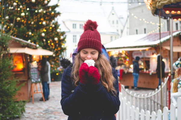 青い冬のコートのハンサムな若い女性 赤い帽子と手袋はココアを楽しんで クリスマスツリーとバックグラウンドのクリスマス運賃で寒い冬の日にタリンタウンホール広場に立っています ストック写真