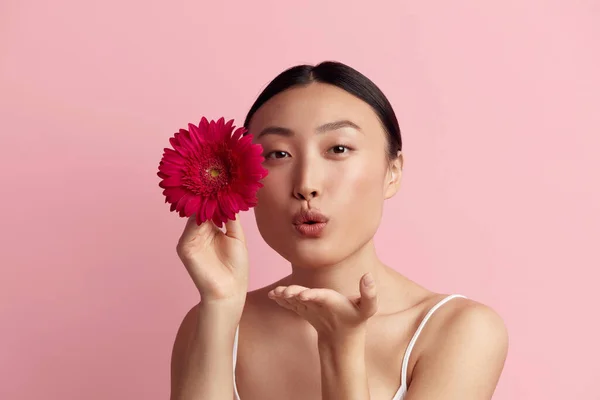 stock image Skin Care Woman Posing with Red Flower. Asian Model Sending Air Kiss Holding Flower. Female With Nude Makeup, Smooth, Hydrated And Glowing Skin. Natural Beauty On Pink Background 