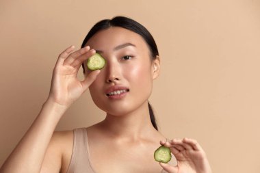 Salatalık tutan Asyalı kadın. Güzel Kız Salatalık Dilimi 'nin Portresi. Gülümseyen genç kadın kameraya bakıyor. Skincare kavramı. Bej arka planda izole edilmiş. Stüdyo Çekimi 