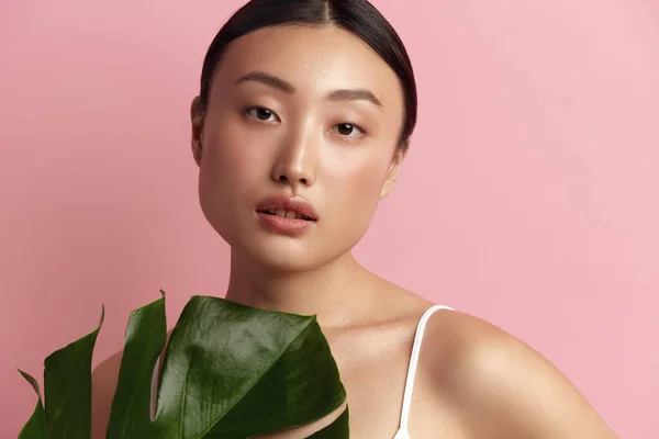 stock image Skin Care Woman Posing with Green Leaf. Asian Model Holding Tropical Plant and Looking At Camera. Female With Nude Makeup, Smooth, Hydrated And Glowing Skin. Natural Beauty On Pink Background 