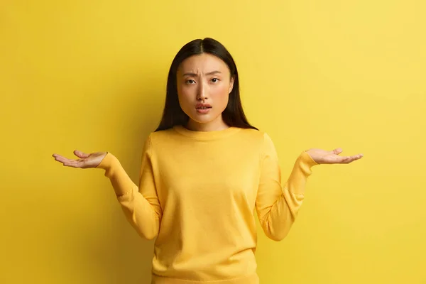 Confused Woman Shrugging Hands. Portrait of Uncertain Asian Girl Expressing Doubts and Bewilderment, Looking at Camera with Question So What, Who Cares. Indoor Studio Shot Yellow Background
