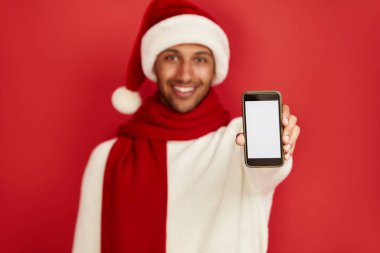 Akıllı Telefon Kullanan Mutlu Adam. Santa Hat 'teki çok ırklı adam cep telefonunu gösterirken gülümsüyor, sosyal medyada sohbet ediyor, mobil servisin tadını çıkarıyor. Reklam için Boş Kopya Alanı, Salon Stüdyosu Çekimi 