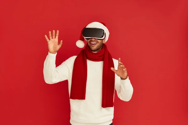 stock image Focused Man Trying VR. Portrait of Multiracial Amazed Guy in Santa Hat Discovering New Technologies Wearing Virtual Reality Headset, Futuristic 3d Vision. Indoor Studio Shot Isolated Red Background 