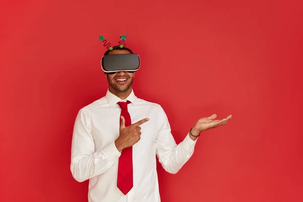 VR Smiling Man Pointing Hand. Positive Multiracial Guy in Virtual Reality Glasses Pointing Away Paying Your Attention at Empty Space for Advertisement. Indoor Studio Shot Isolated on Red Background