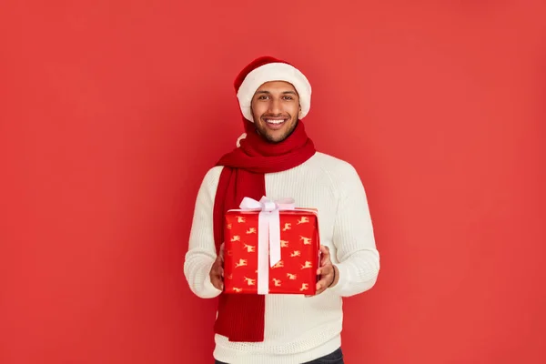 Happy Man Holding Present. Excited Multiracial Guy in Santa Hat Holding in Hands Red Wrapped Present Box with Excitement. Indoor Studio Shot Red Background