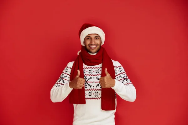 Happy Man Thumbs Up. Pleased Multiracial Man in Santa Hat Smiling to Camera and Gesturing Thumbs Up, Approval Hand Sign, Satisfied with Result. Indoor Studio Shot Isolated on Red Background