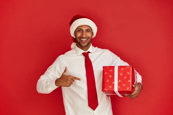 Man Pointing New Year Present. Multiracial Guy in Santa Hat Paying Attention at Gift Box and Looking at Camera. Red Background, Studio Shot