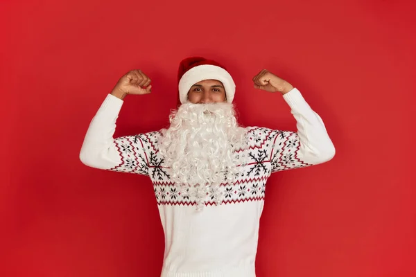 Santa Claus Showing Biceps. Portrait of Confident Proud Multiracial Man Showing Biceps, Feeling Power Success. Indoor Studio Shot Isolated on Red Background