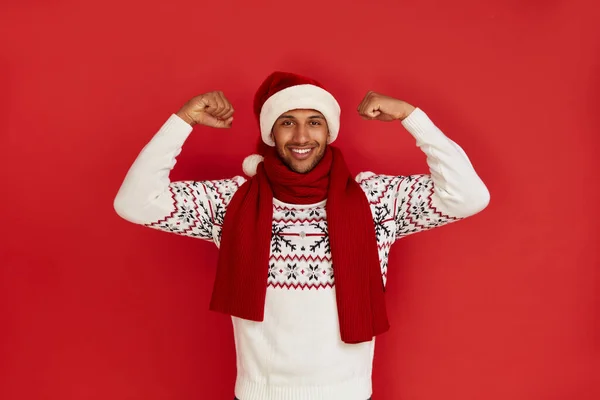 Multiracial Man Showing Biceps. Portrait of Confident Proud Man in Santa Hat Showing Biceps, Feeling Power Success. Indoor Studio Shot Isolated on Red Background