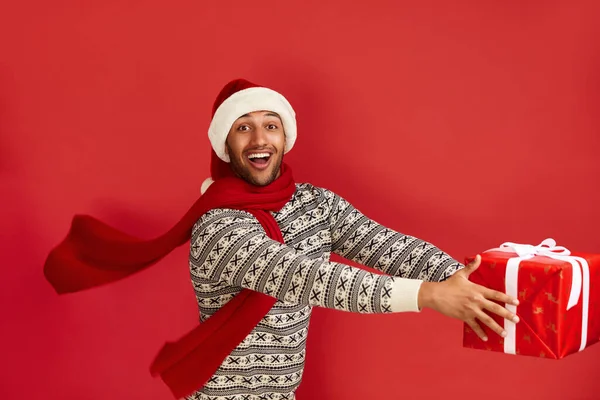 Happy Man Holding Present. Excited Multiracial Guy in Santa Hat Holding in Hands Red Wrapped Present Box with Excitement. Indoor Studio Shot Red Background