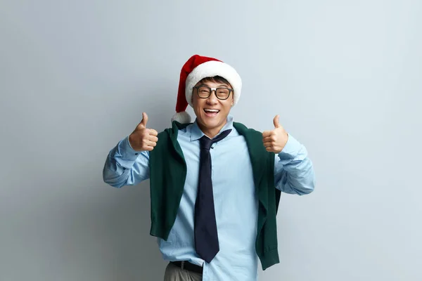 Happy Man Thumbs Up. Pleased Asian Man in Santa Hat Smiling to Camera and Gesturing Thumbs Up, Approval Hand Sign, Satisfied with Result. Indoor Studio Shot Isolated on Grey Background