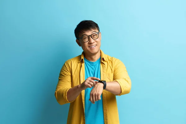 Asian Man Pointing Watch. Portrait of Cheerful Punctual Guy Pointing at Wrist Watch and Smiling, Showing Smartwatch Devise with Mock Up Display. Indoor Isolated on Blue Background