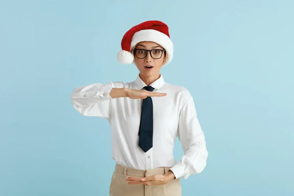 Surprised Businesswoman Showing Big Size. Satisfied Caucasian Lady in Santa Hat Showing Size With Both Hands, Looking at Camera With Surprised Expression. Indoor Shot Isolated on Blue Background