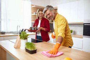 Gülümseyen Adam Temizlik Masası. Ahşap Mutfak Masası 'ndan kıdemli adam kırıntılarını siliyor. Temizlik için Erkek Temizlik Mendili Kullanan Olgun. Eşi Mutlu Yüzle Duruyor