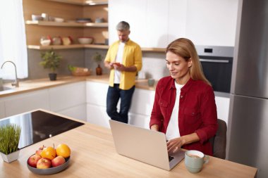 Mutfakta dizüstü bilgisayar kullanan sarışın kadın. Mutlu çiftler hafta sonlarını evde birlikte geçiriyorlar. Gri saçlı adam arka planda akıllı telefona bakıyor. 