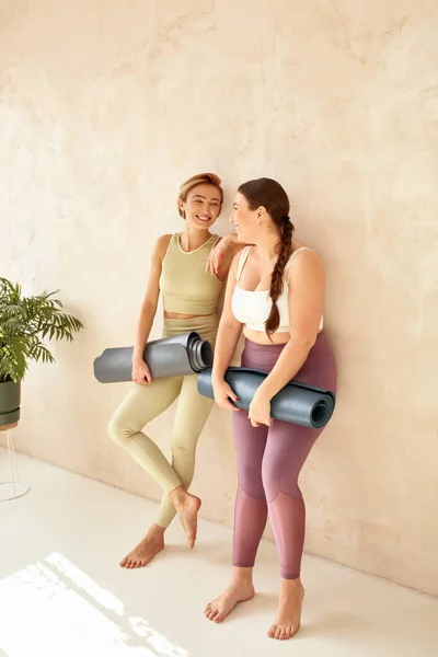 Women Embracing After Yoga. Female Friends Laughing And Holding Yoga Mats After Yoga Session Together At Home. Attractive Girls In Sportswear Spending Free Leisure Time
