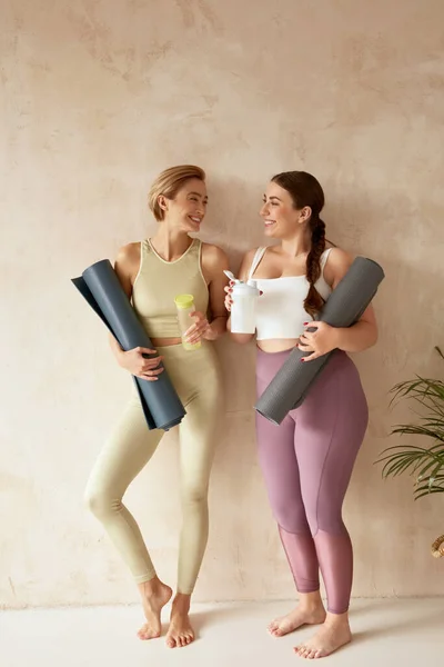 Smiling Woman Talking After Yoga. Female Friends Laughing And Holding Detox Drinks After Yoga Session Together At Home. Attractive Girls In Sportswear Spending Free Leisure Time