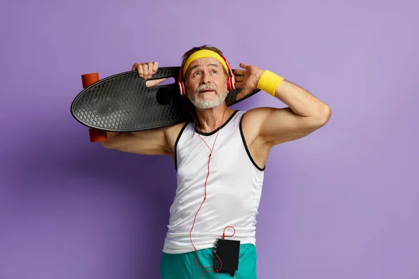 微笑的男人拿着滑板 头戴耳机的老年人在背负和聆听音乐的同时 在紫罗兰背景下被隔离 — 图库照片