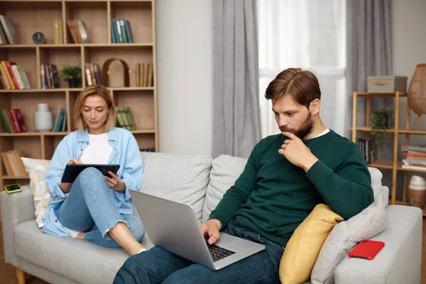 Couple Using Gadgets At Home. Married Couple Of Freelancers Work From Home. Guy And Girl In Casual Clothes With Gadgets Work In The Living Room At The Sofa