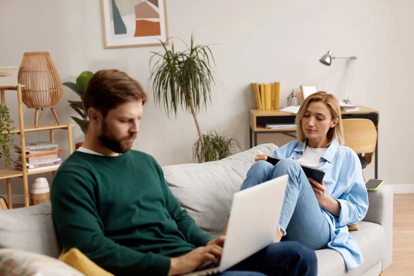 Couple Using Gadgets At Home. Married Couple Of Freelancers Work From Home. Guy And Girl In Casual Clothes With Gadgets Work In The Living Room At The Sofa